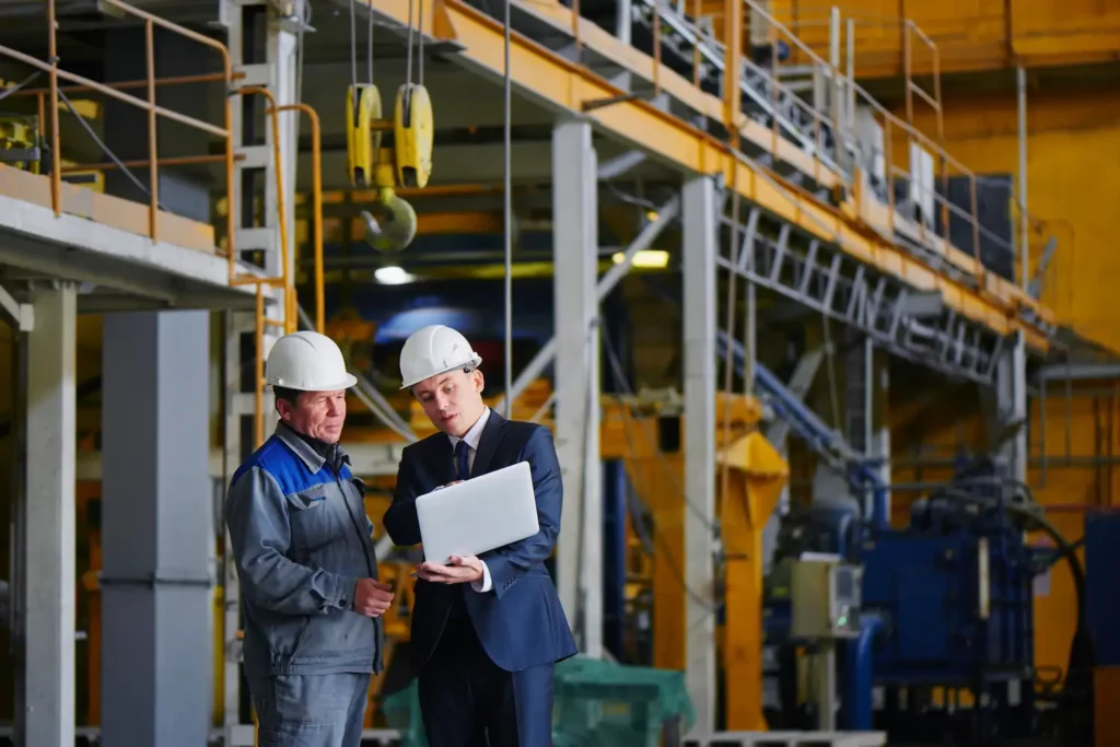 Der Mann im Anzug und mit Helm hält den tragbaren Computer in der Hand und zeigt ihn auf dem Bildschirm dem Arbeiter im Overall in einem Industriegebäude